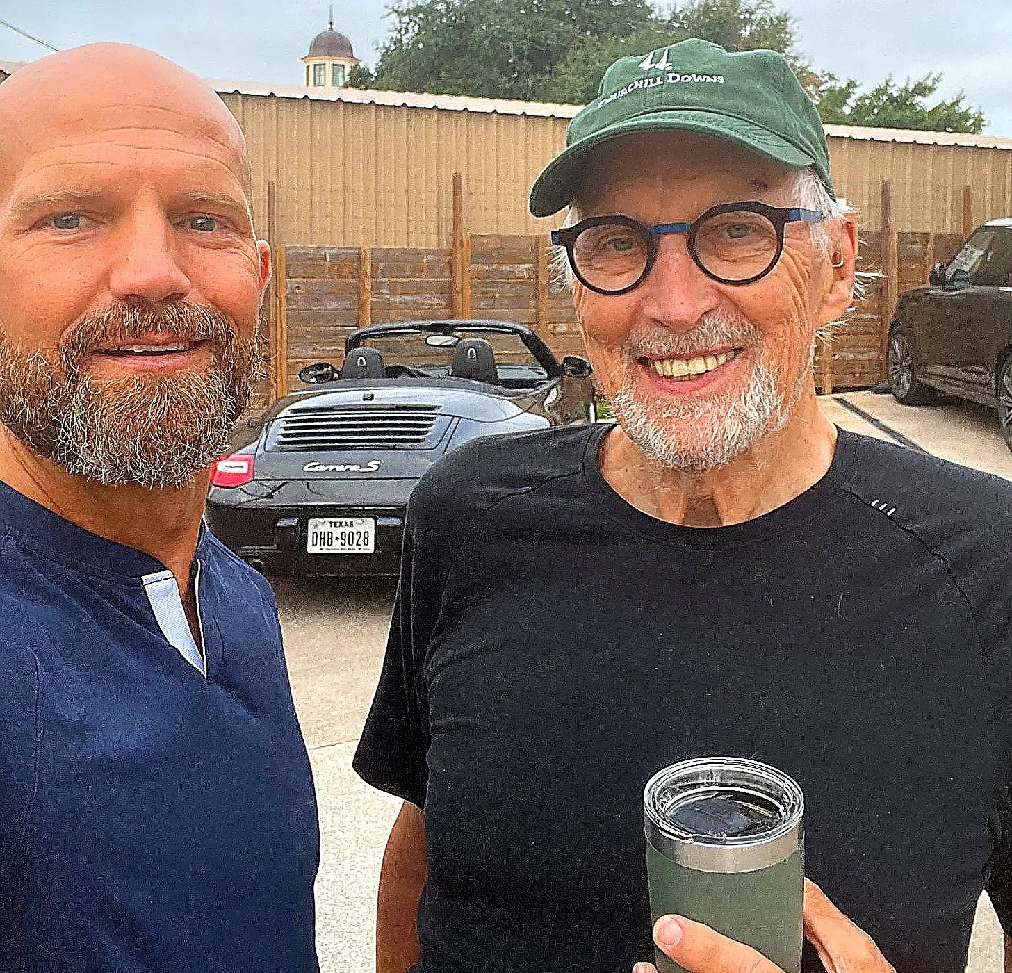 Outdoor selfie of two smiling bearded men standing beside a convertible sports car; the man on the right wears glasses and holds a green insulated cup.